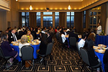 A room filled with round tables, covered in blue table cloths. People are paying attention to an individual speaking at the front of the room. 